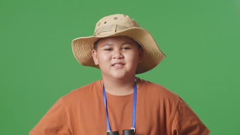 niño turista asiático con un sombrero y binoculares sonriendo a la cámara mientras está de pie con los brazos akimbo en el fondo de la pantalla verde. niño investigador examina algo, aventura de turismo de viaje, de cerca