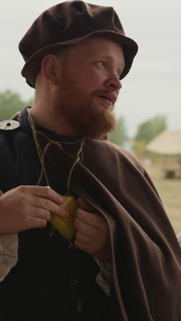 bearded man in medieval clothing talks standing in yard in countryside. village squire holds yellow potion bottle in bosom closeup on blurred background