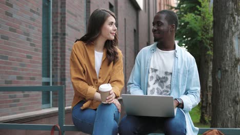 cámara haciendo zoom en un hombre afroamericano y una mujer caucásica usando una laptop y sosteniendo una taza de café sentada en la calle cerca de la universidad
