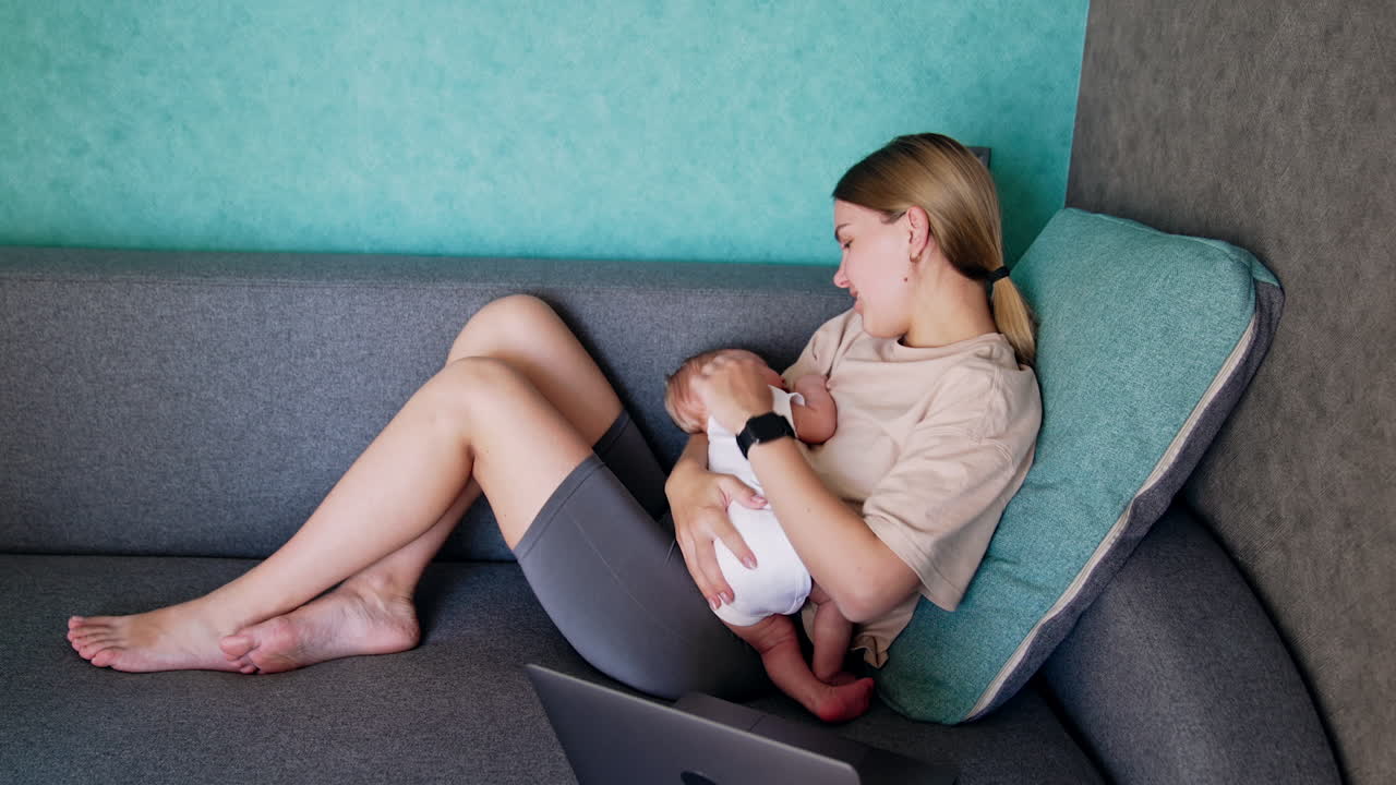 Happy mom sits comfortably on the sofa with newborn in hands. Woman strokes baby head tenderly and then turns to a laptop.