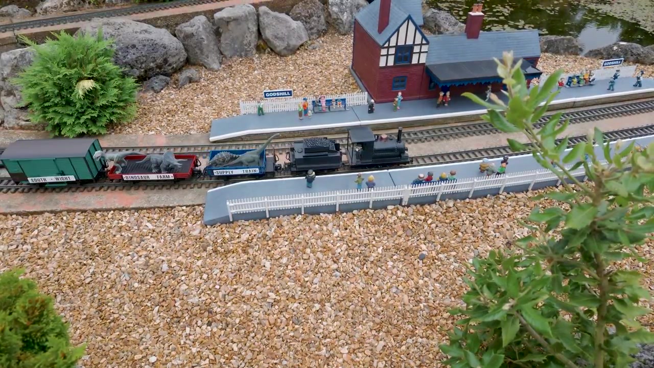 Modeltog ankommer til Godshill Model Village station, kører ind og stopper, mens dinosaur-tema vogne passerer miniatureperroner, små figurer og bygninger i en britisk attraktion.