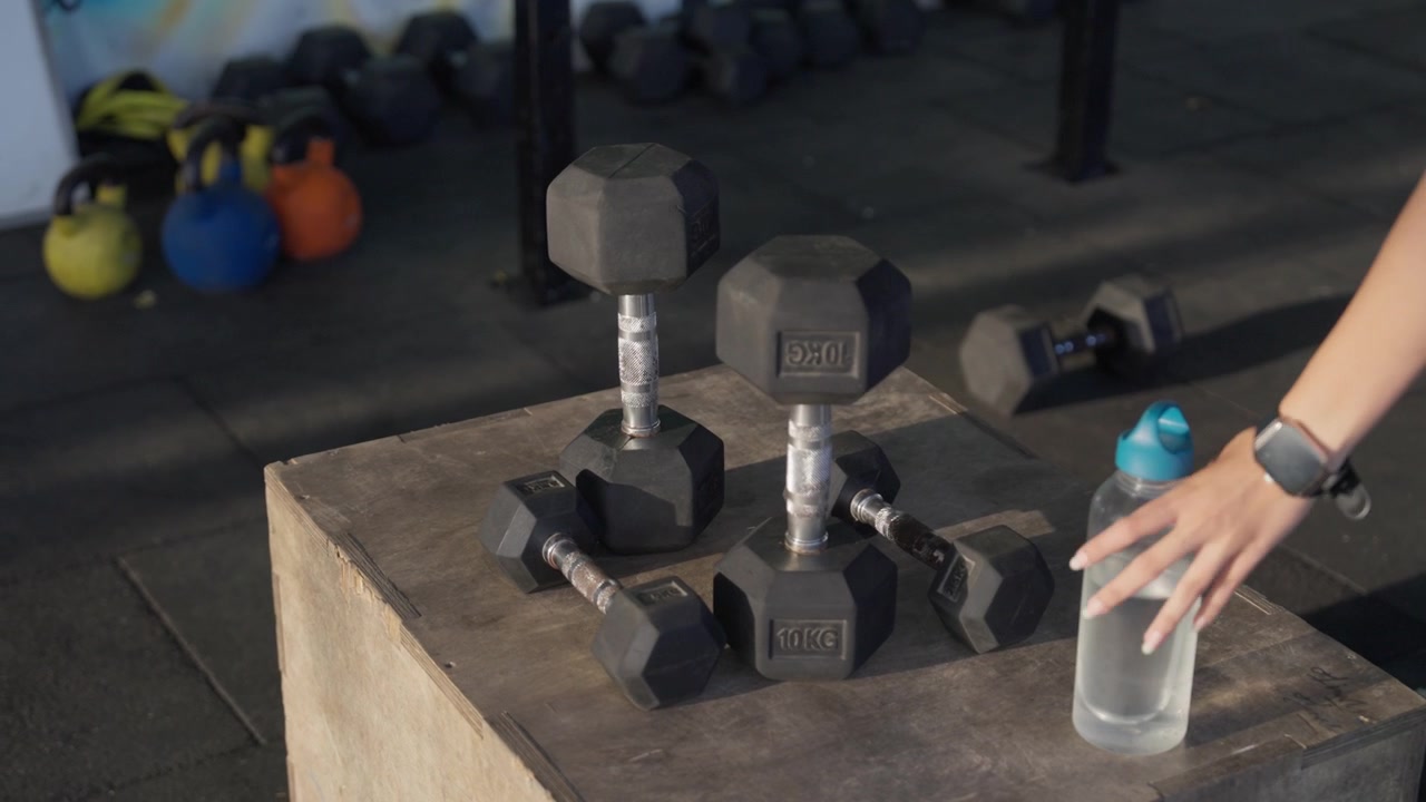 Woman wearing a watch reaching for and picking up a water bottle placed beside hex dumbbells on a wooden plyometric box in a modern indoor gym.