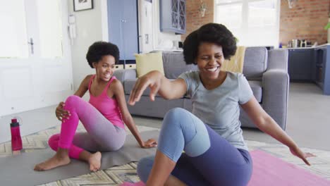 Happy-african-american-mother-and-daughter-doing-yoga-in-living-room,-stretching