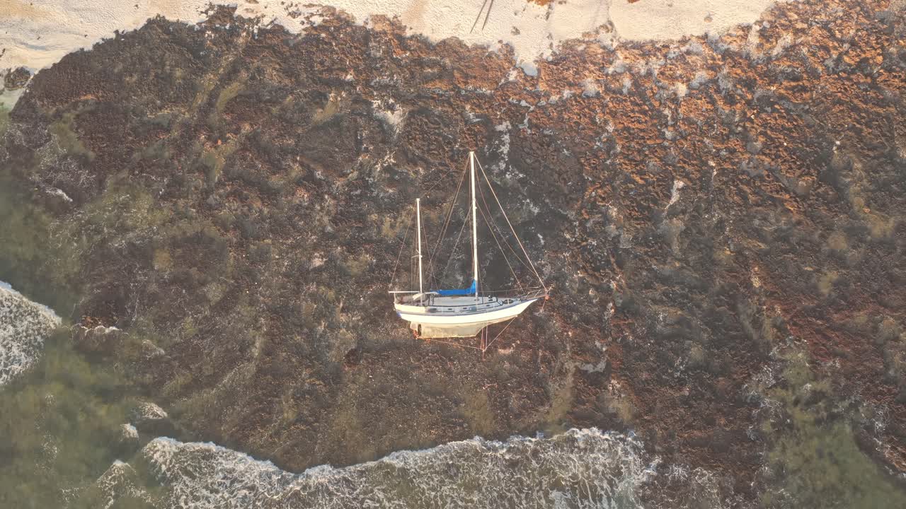 Stranded Sailboat tilting against rocky shoreline of Playa del Medio Beach, Fuerteventura, with crashing ocean waves under bright sunlight, top down ascending shot