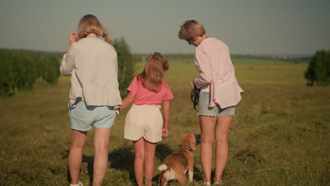 little girl touches dog's head while her aunt feeds dog, with her mother adjusting her hair beside them in vast green field under sunny sky, with there hair and cloth blown by the wind