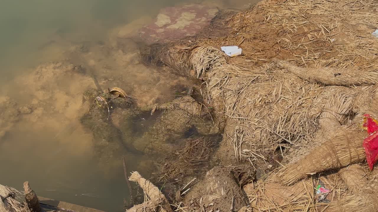 Murky river water flows through scattered idol remnants and straw frameworks along the bank, mixing silt and debris as it moves through the submerged materials