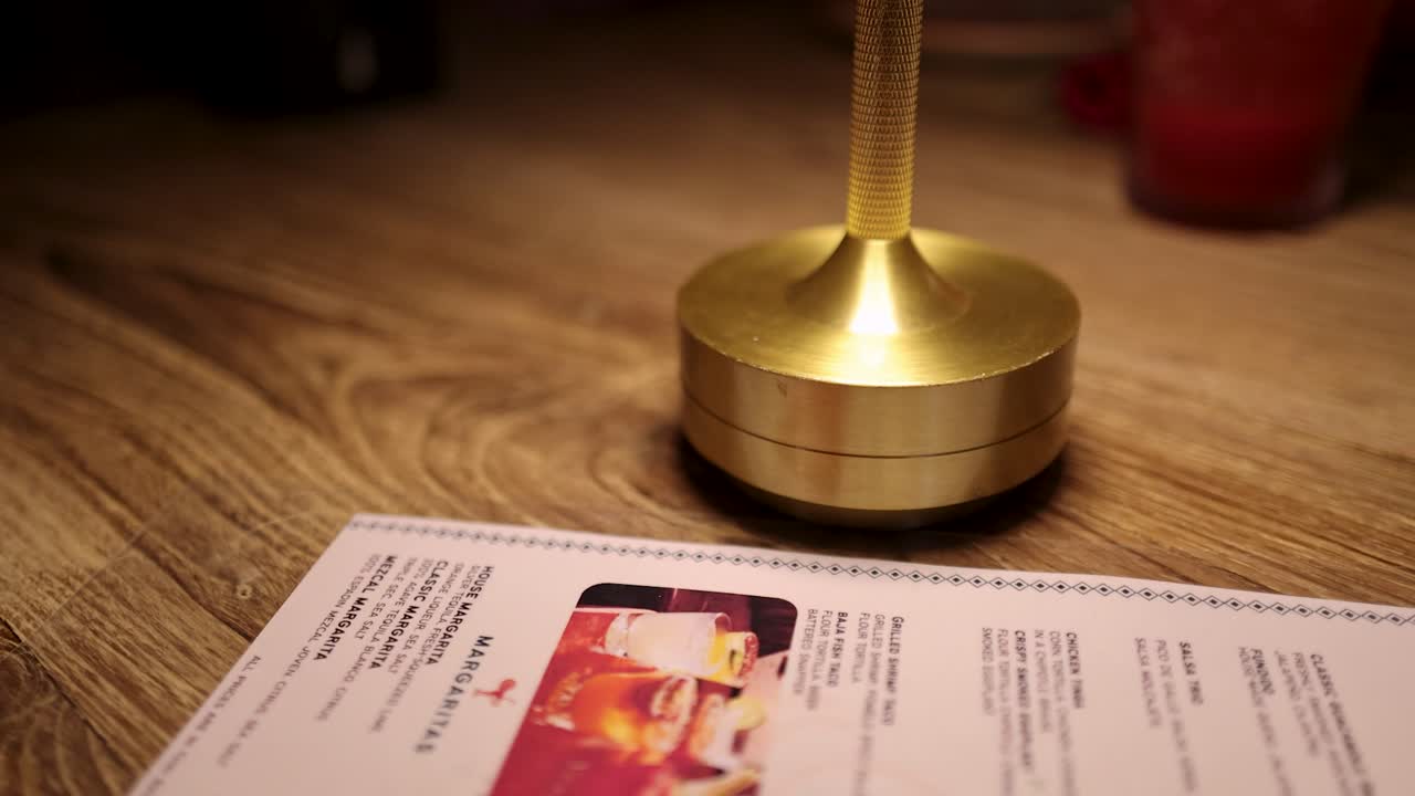 A close-up of a gold lamp base and menu on a wooden restaurant table under warm lighting, with drinks blurred in the background. Minimal camera movement