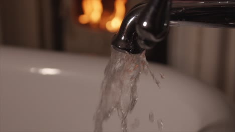 Close-up-of-running-bath-tap-with-fireplace-in-background