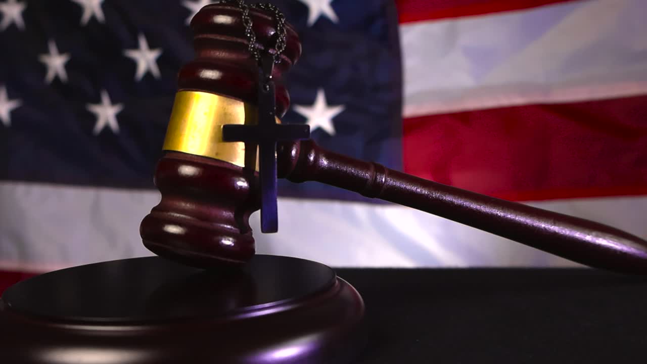 Wooden court justice gavel resting on a wooden dark desk with a holy christianity cross hanging on the bronze decorations and a American United states flag in the bokeh blurry background
