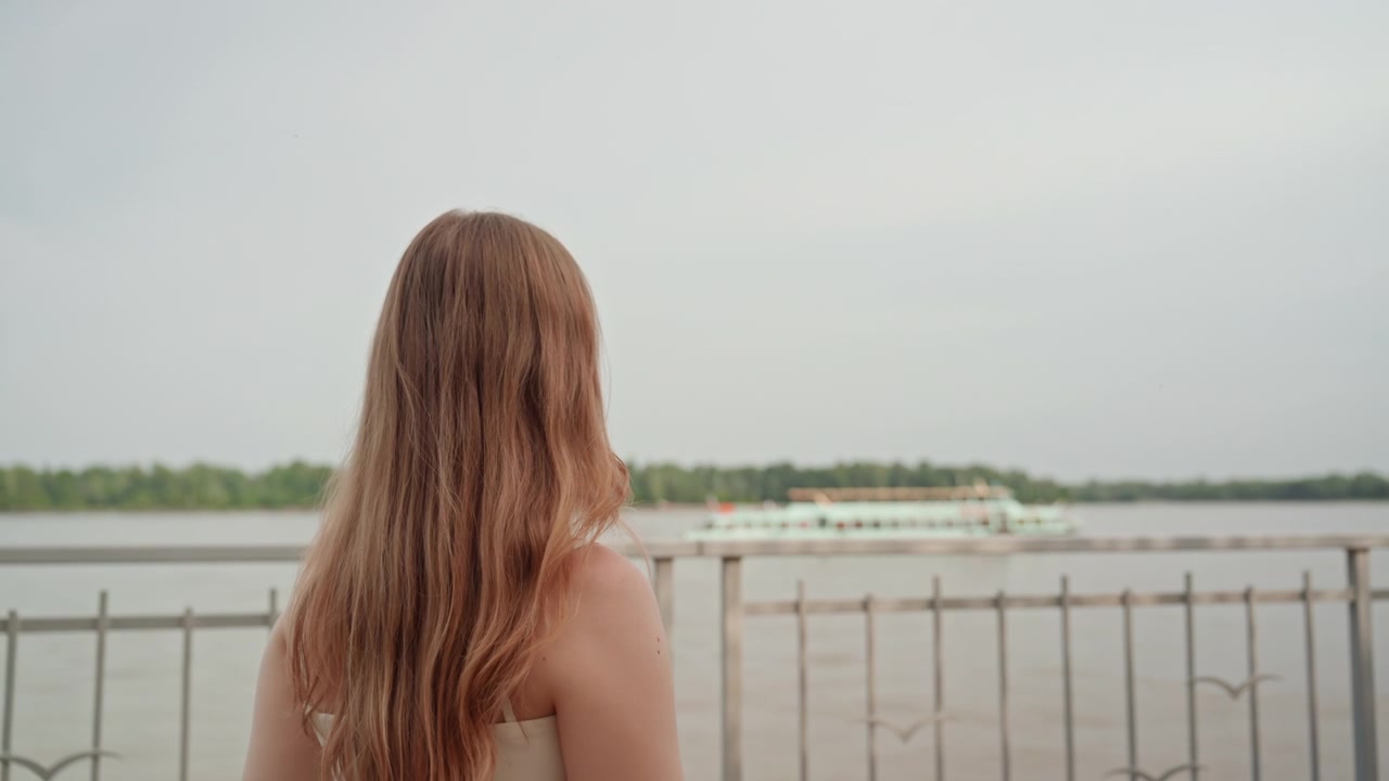 Woman with long hair stands by riverside railing facing away from camera and looking at water. Woman wears light colored clothing as boats pass in background under overcast sky with trees