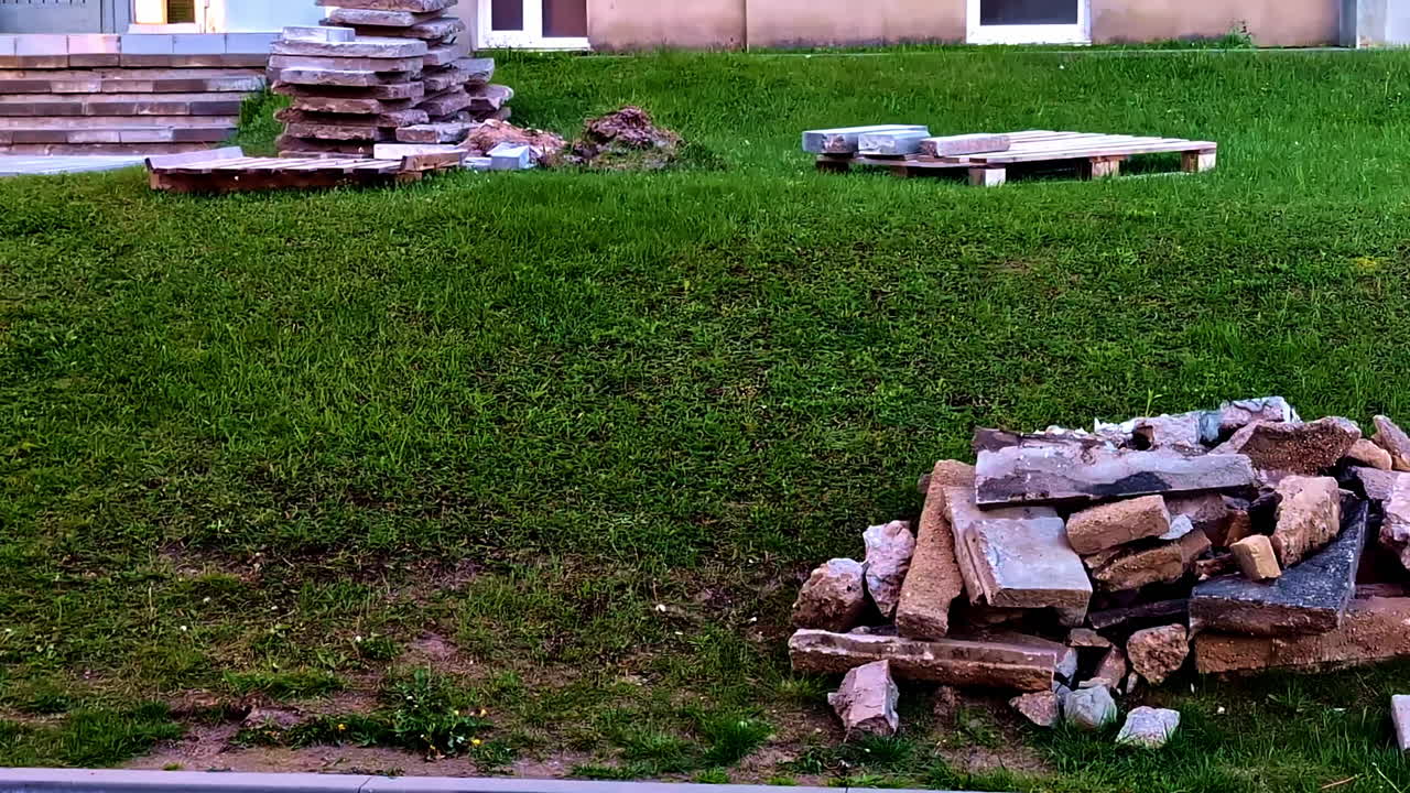 Broken paving stones and construction debris lie scattered on green grass near a building