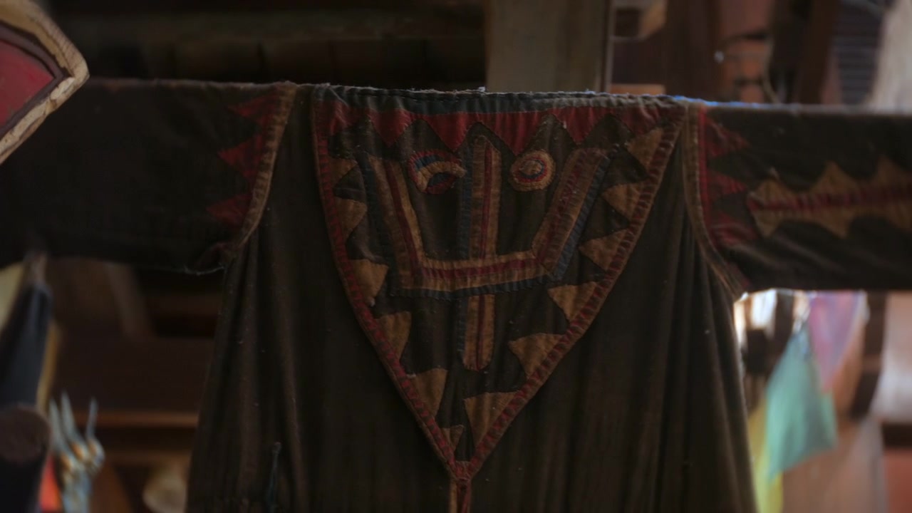Abiti in tessuto appesi all'interno di un santuario tibetano, che mostrano un tessuto nero usurato con motivi cuciti rossi e marrone chiaro. La vista ravvicinata evidenzia la decorazione rituale fatta a mano, la luce interna soffusa e la texture invecchiata.