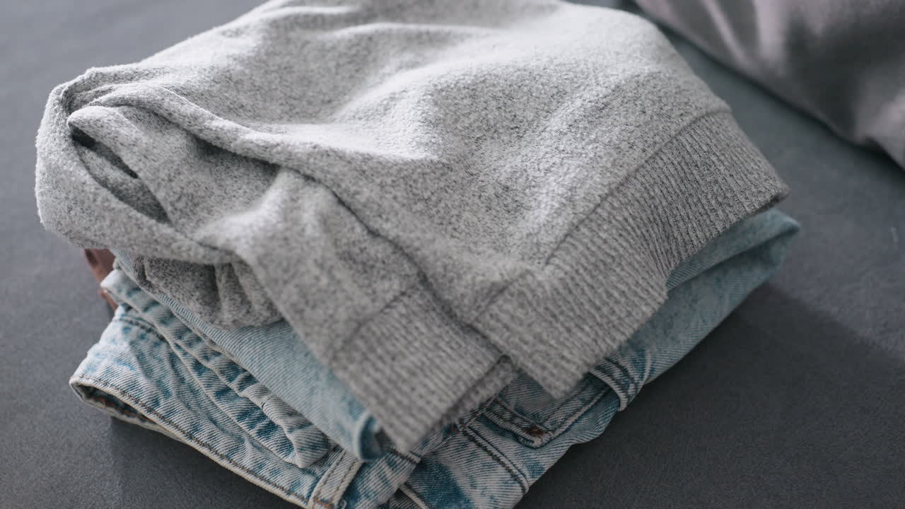 Close up of housekeeper placing neatly folded jeans on top of brown cloth stacked on couch showing detail of domestic clothing care household organization lifestyle responsibility