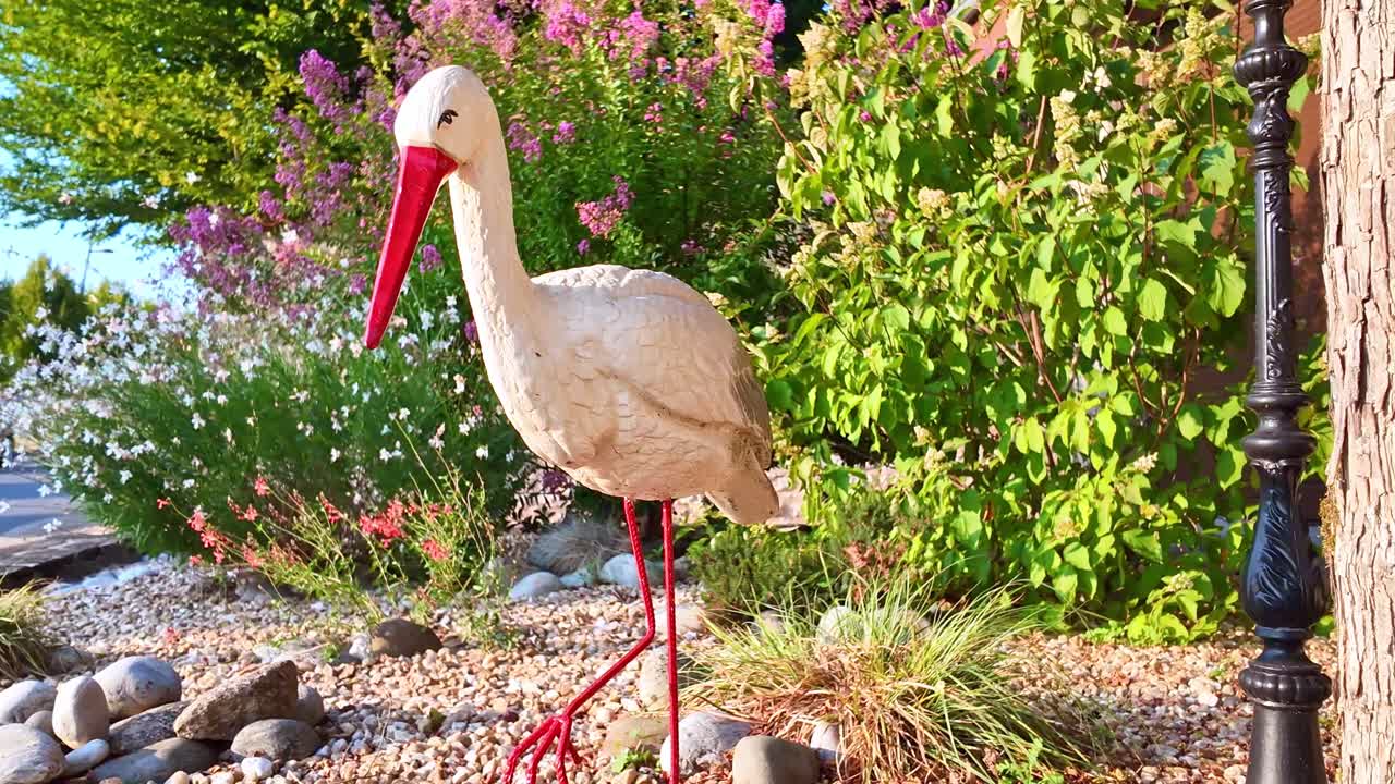 Statue of a white stork standing in garden with red beak, lush background, daylight, medium approach