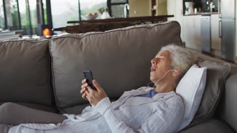 Senior-mixed-race-woman-lying-on-sofa-wearing-earphones-and-using-smartphone