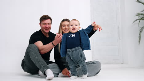 happy smiling baby boy stands supported by his parents. loving couple sit on the floor with their beloved child. white backdrop.