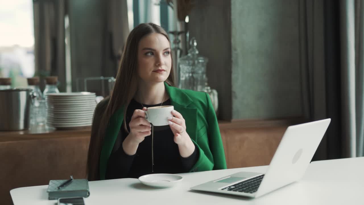 bela jovem sentada em um restaurante elegante vestida com trajes de negócios bebe café e olha contemplativamente pela janela enquanto está sentada atrás de seu laptop
