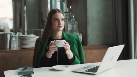 beautiful young woman sitting in a stylish restaurant dressed in business attire sips coffee and contemplatively gazes out the window while sitting behind her laptop