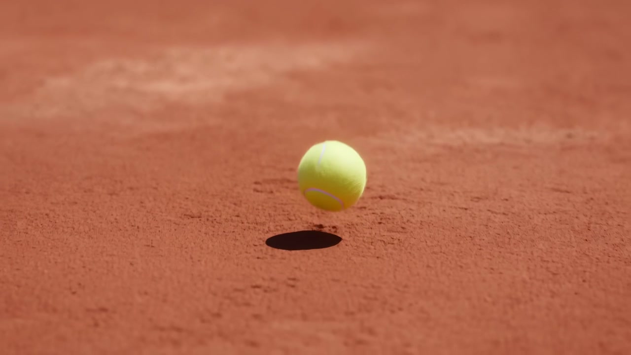 Kirkkaan keltainen tennispallo nähtynä ilmassa punaisen massakentän yllä lähellä takarajaa, voimakas ylävalo luo terävän varjon ja selkeän erottelun pinnasta, vangiten energisen ulkopelin.