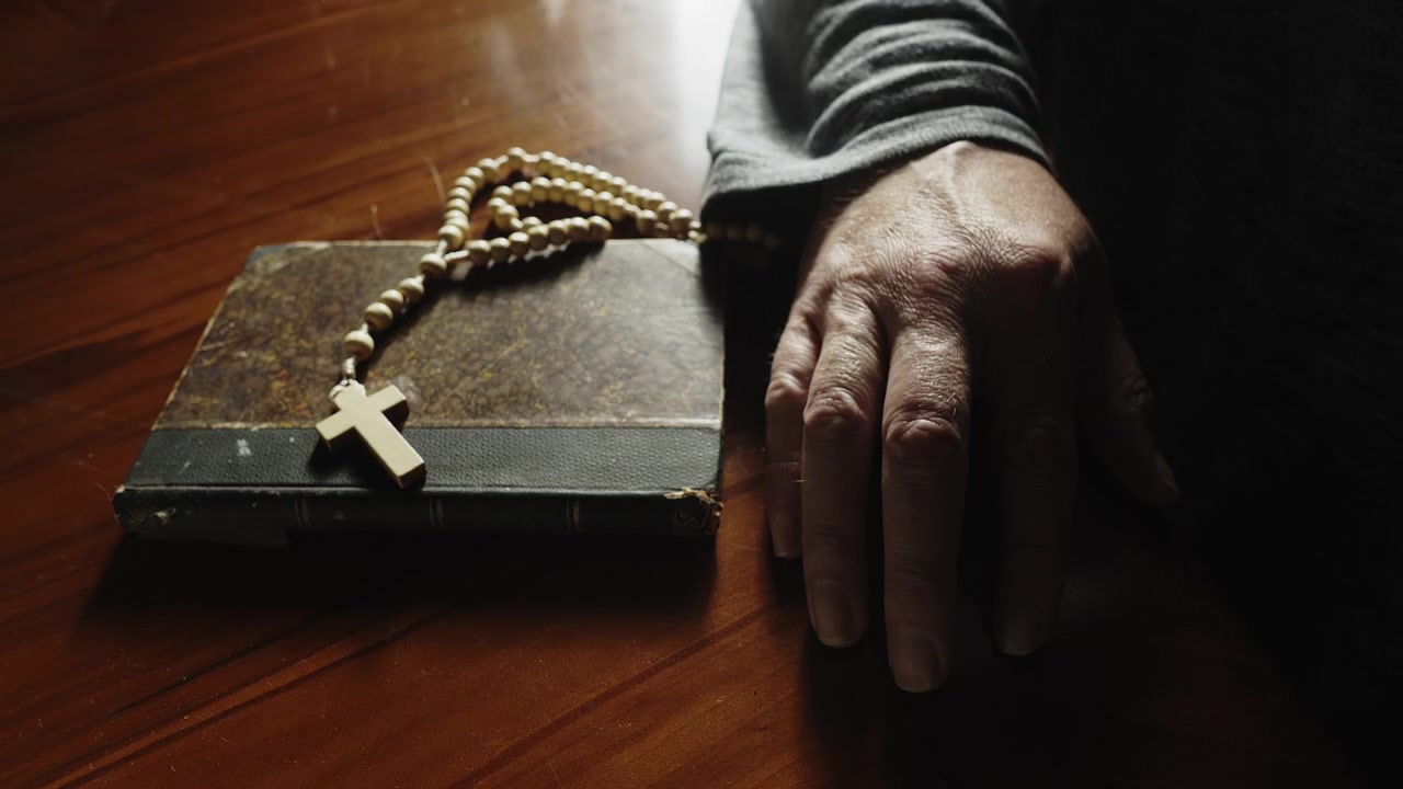 Primo piano di mani che avvicinano un libro di preghiere e un rosario prima di allontanarli, raffigurando conflitto spirituale, dubbio, turbolenza interiore e una crisi di fede personale