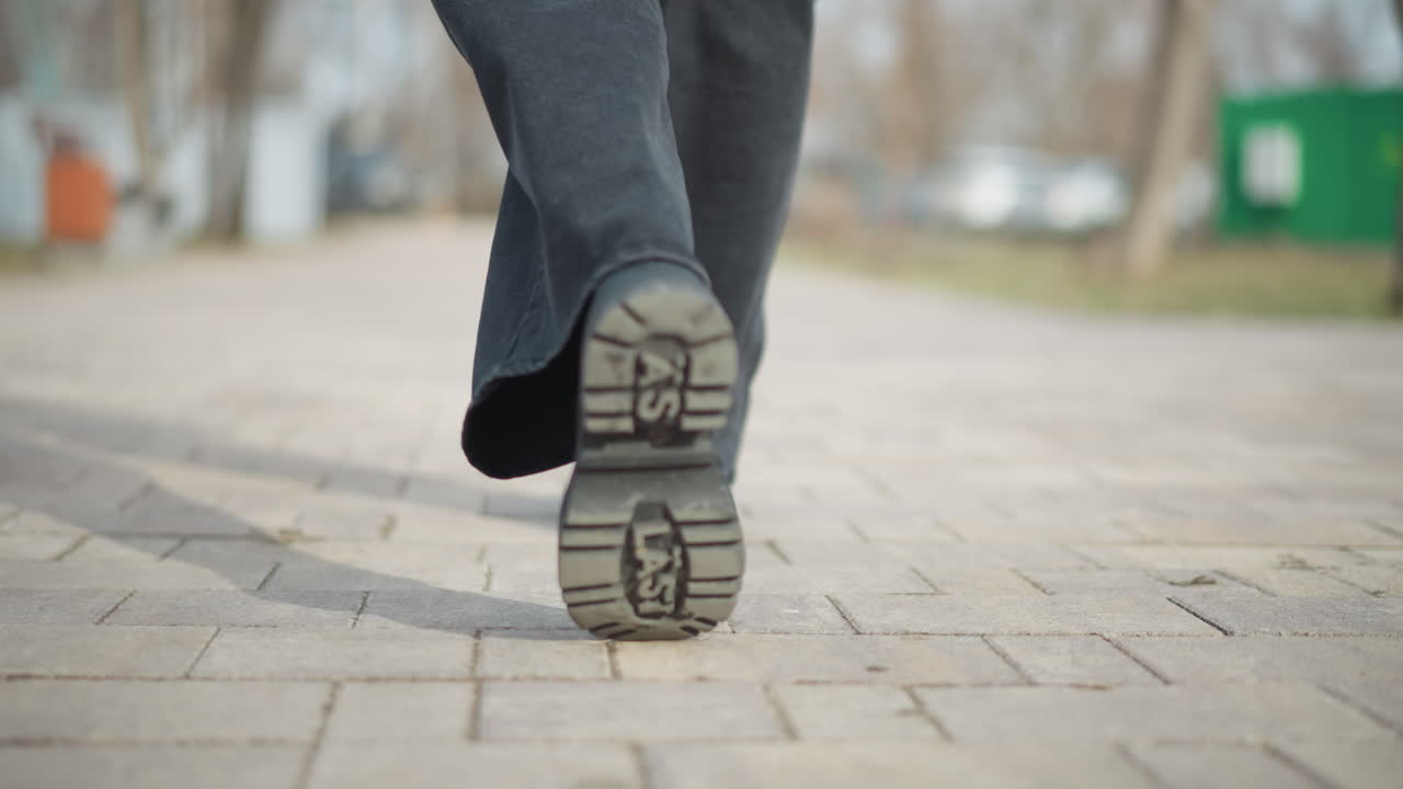 Low-angle close-up captures bottom of raised boot as woman walks forward on paved park path, emphasizing movement, footwear tread design, and urban outdoor setting in daylight with soft background