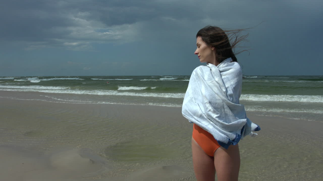 Frozen wet beautiful woman in a swimsuit wrapped with a cold towel to keep warm. Stands alone on the sea beach during windy weather