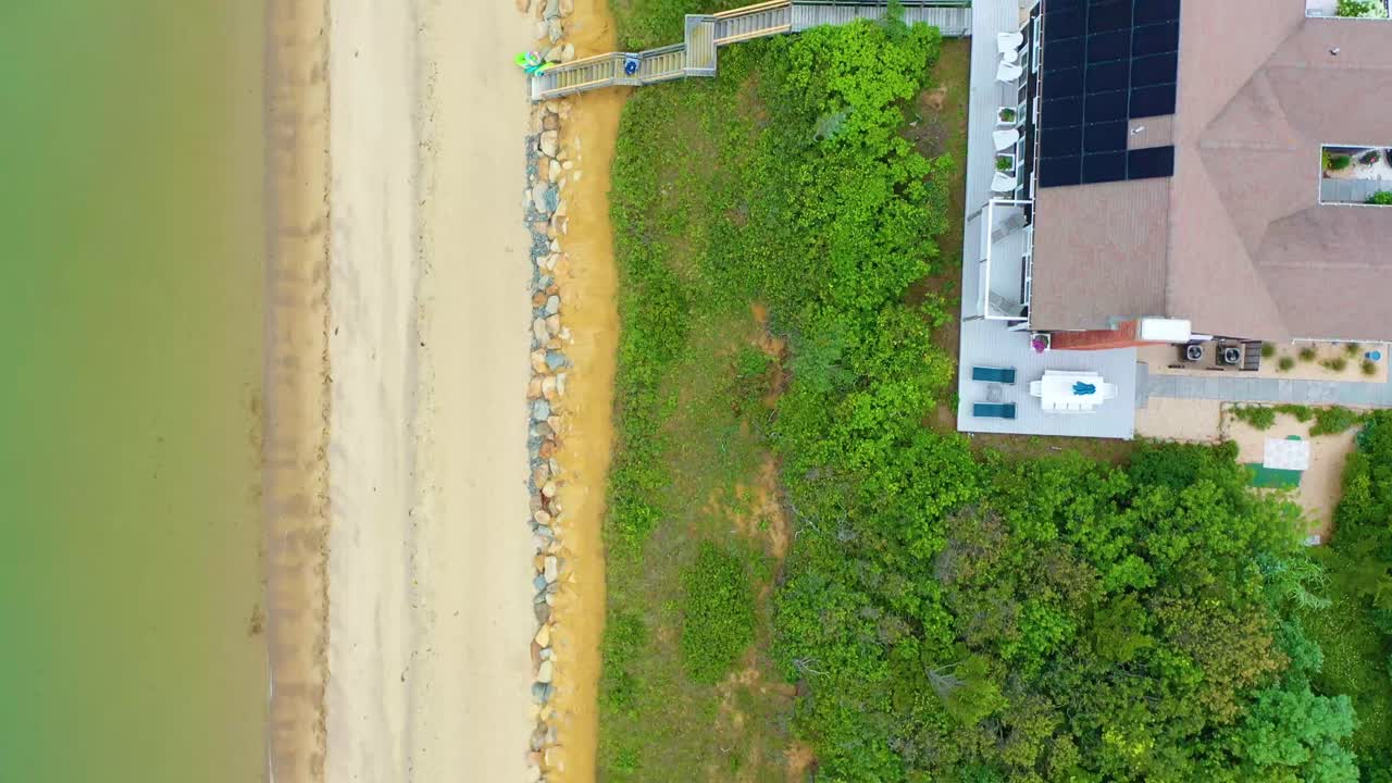 Bird’s-eye drone shot of coastal neighborhood with houses, sandy beach, and calm waters, framed by dense green trees and paths leading from homes directly to the shoreline