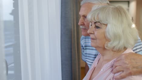 Una-Encantadora-Pareja-De-Ancianos-Maduros-Se-Abraza,-Se-Abraza-Y-Mira-Por-La-Ventana-De-Su-Casa.