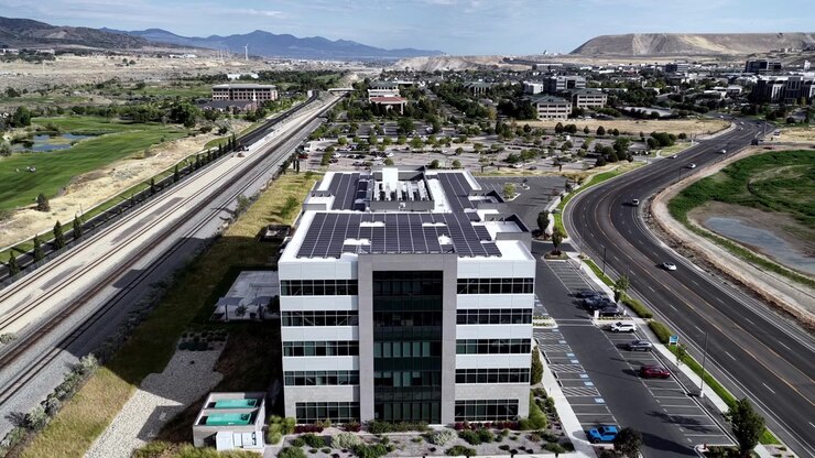 Un drone in allontanamento rivela un moderno edificio per uffici a Lehi, Utah, alimentato da pannelli solari sul tetto, circondato da montagne, parcheggi e strade moderne in una giornata limpida e soleggiata. Nessuna persona presente.