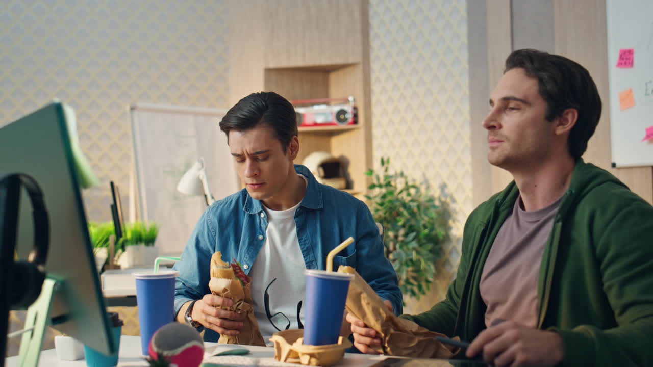 샌드위치를 먹는 사업가들이 가까이에서 이야기하고 있습니다. 컴퓨터를 쳐다보는 사람들