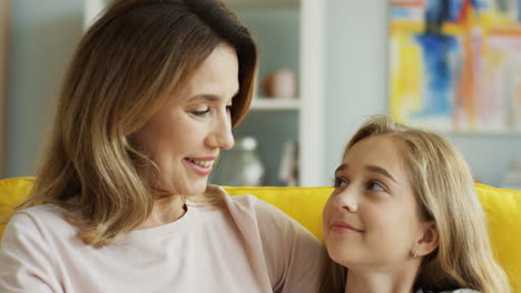 cerca de una madre e hija felices y sonrientes sentadas en un sofá amarillo y mirándose con amor