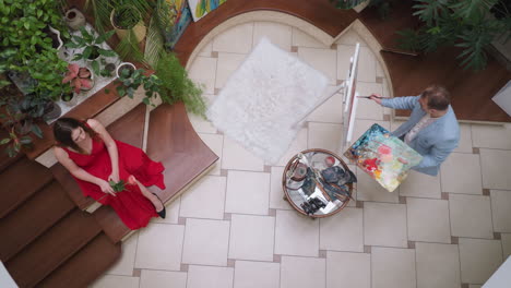 mujer con vestido escarlata y artista en el caballete. retratista dibuja a una mujer atractiva con una flor sosteniendo la paleta de colores en la galería. proceso de creación de arte inspirador