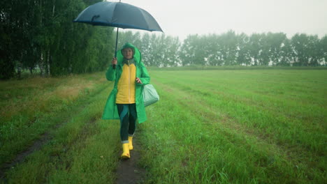 woman in green raincoat putting up her black umbrella while walking along a path surrounded by greenery, she is wearing boots and carrying a bag, with trees in the background under a misty sky