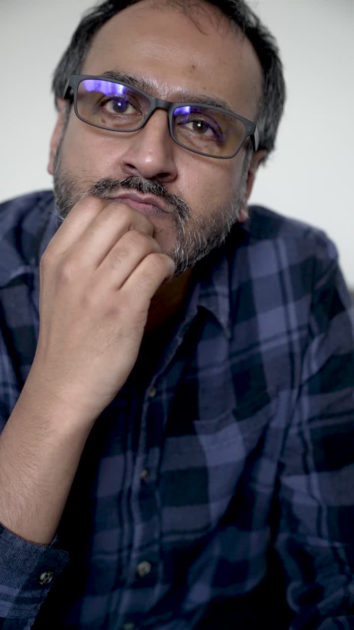 South asian man with beard and glasses touching his chin, contemplating. vertical video