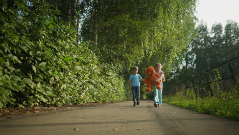 two kids walking along tree lined paved path, girl carrying large teddy bear while boy walks beside her, afternoon sunlight highlights their journey home, surrounded by nature and iron fencing