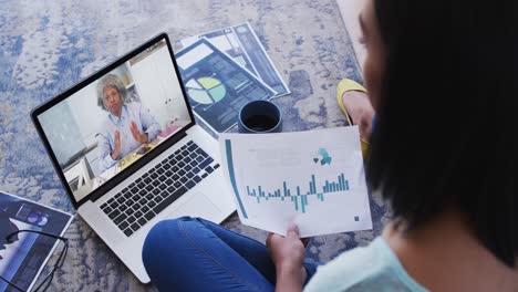 Mixed-race-businesswoman-sitting-on-floor-using-laptop-having-video-call-with-female-colleague