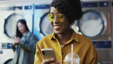 Girl-Drinking-Orange-Juice-With-Straw,-Texting-Message-On-Phone-And-Waiting-For-Clothes-To-Get-Clean-Woman-Sipping-Drink-In-Laundry-Service-Room-And-Tapping-Or-Scrolling-On-Smartphone