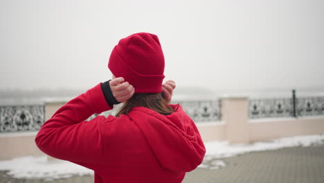 vista trasera de una mujer con capucha roja ajustando su gorra roja al aire libre durante el invierno, entorno urbano con alrededores cubiertos de nieve y barandillas decorativas visibles en el fondo bajo clima de niebla