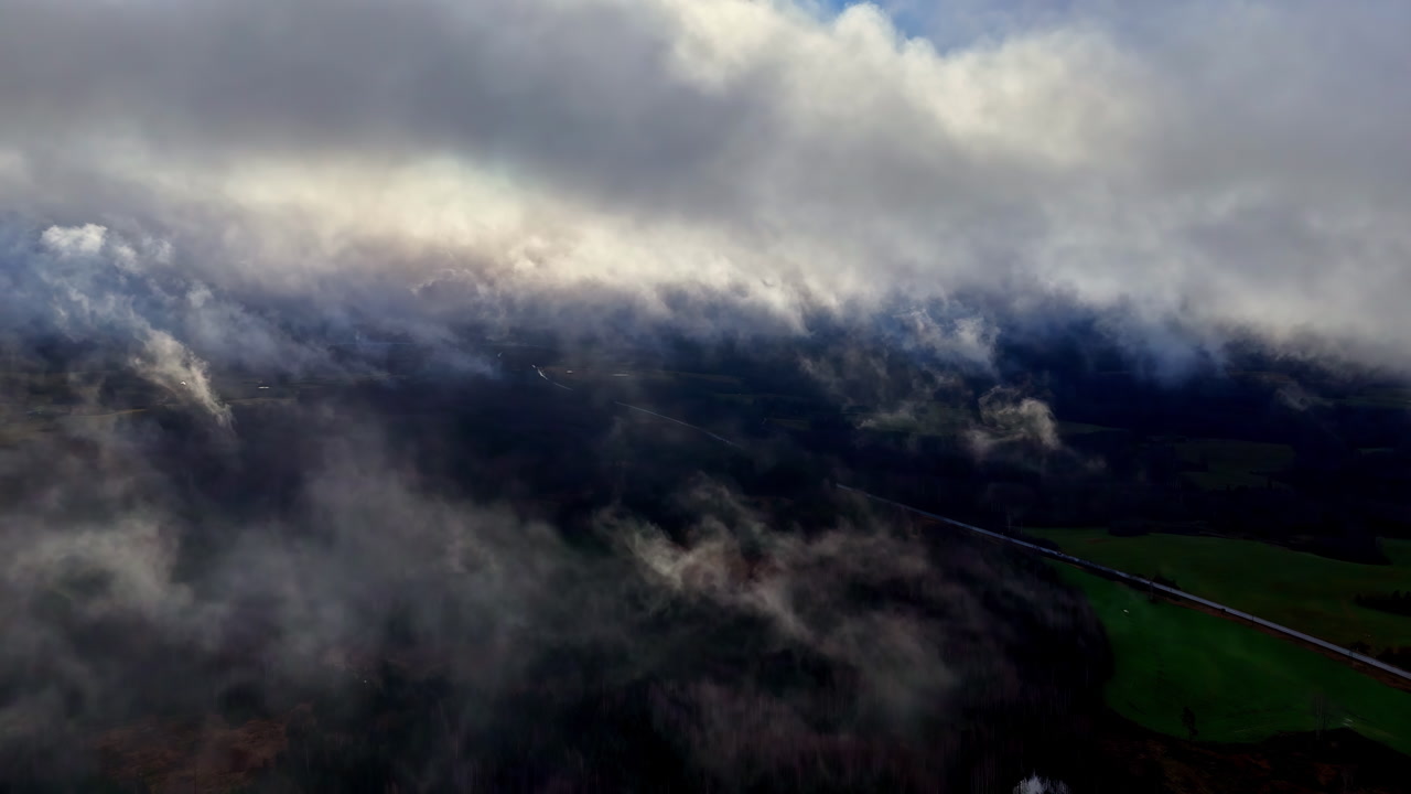 Drone flies through dramatic swirling dark storm clouds above green Latvian countryside with road and fields barely visible beneath turbulent cloudscape