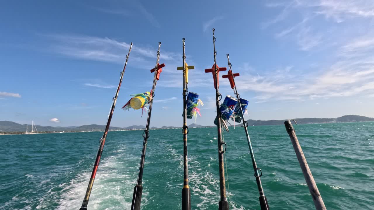 Fishing rods sway on a moving boat under clear skies in Phuket, Thailand. Vibrant ocean and flags create a dynamic scene