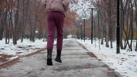primer plano de las piernas y botas de una mujer joven corriendo por un camino cubierto de nieve helada en traje de invierno, en un parque al aire libre con árboles alineados y bayas rojas