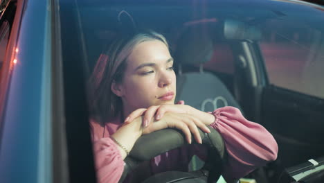 woman in pink dress seen through her car windscreen resting her head on the steering wheel, looking tired, while a car with headlights on drives past in the background during the night