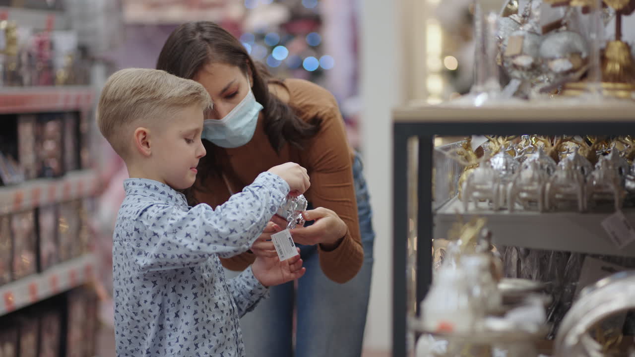 una madre con una máscara médica con su hijo en el centro comercial elige decoraciones caseras para navidad. mira los juguetes del árbol de navidad. epidemia de coronavirus