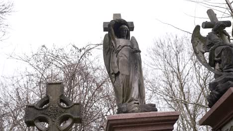 cross and statue on a gravestone covered in moss and green leaves in a forest graveyard on a cloudy day