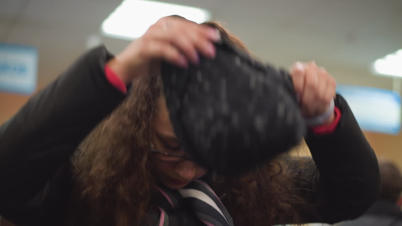 Damsel in glasses wearing black winter jacket and striped scarf adjusting hair indoors, focused expression showing natural candid lifestyle moment with sense of preparation, warmth, comfort atmosphere