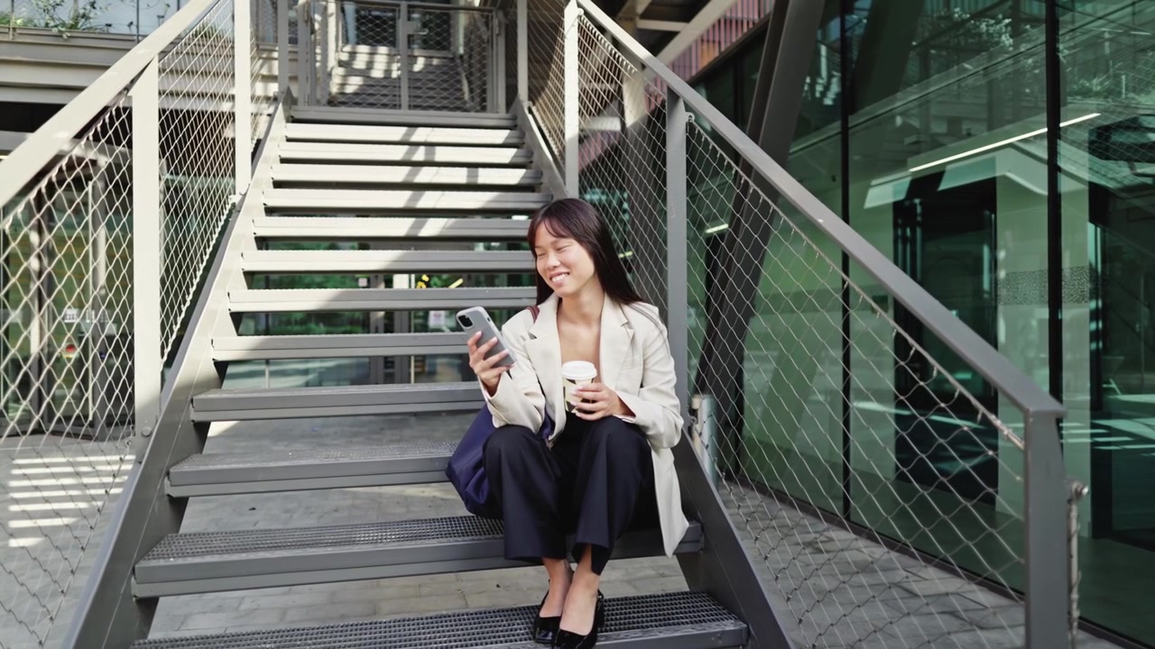 屋外の階段でスマホを使いコーヒーを飲む若いアジア人女性
