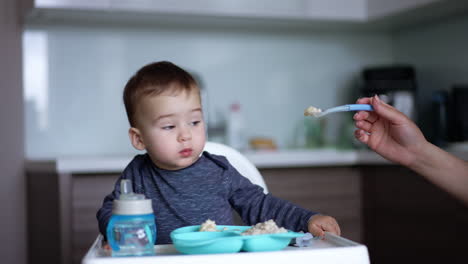 mom shoves a full spoon to her son. little boy refuses to eat and turns away. blurred backdrop.