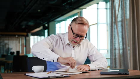 senior man in white shirt pulls the pages out of his paper book. man rolls the papers in a ball and throws them backwards.