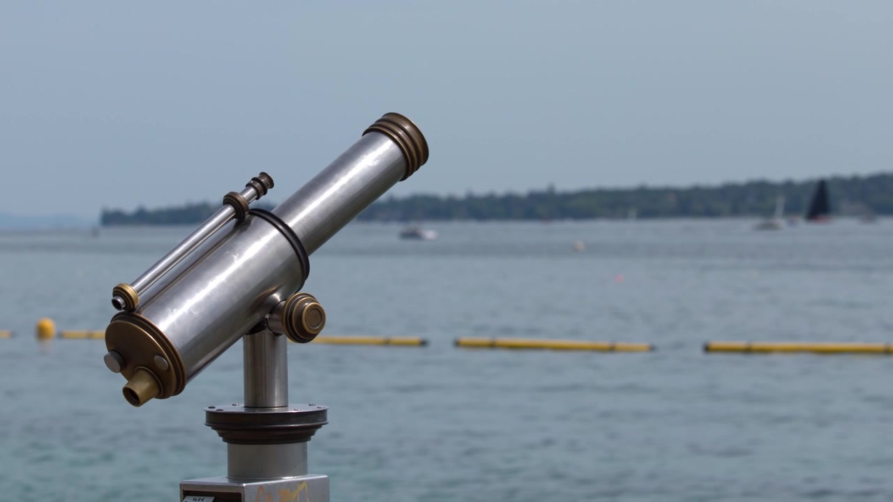Plano médio estático de um telescópio metálico operado por moedas virado para um lago calmo sob luz diurna brilhante.