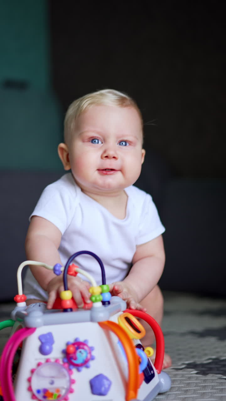 Adorable plump round-faced baby with blond hair plays with a toy. Kid pushes the toy by the floor smiling to the camera adorably. Vertical video.
